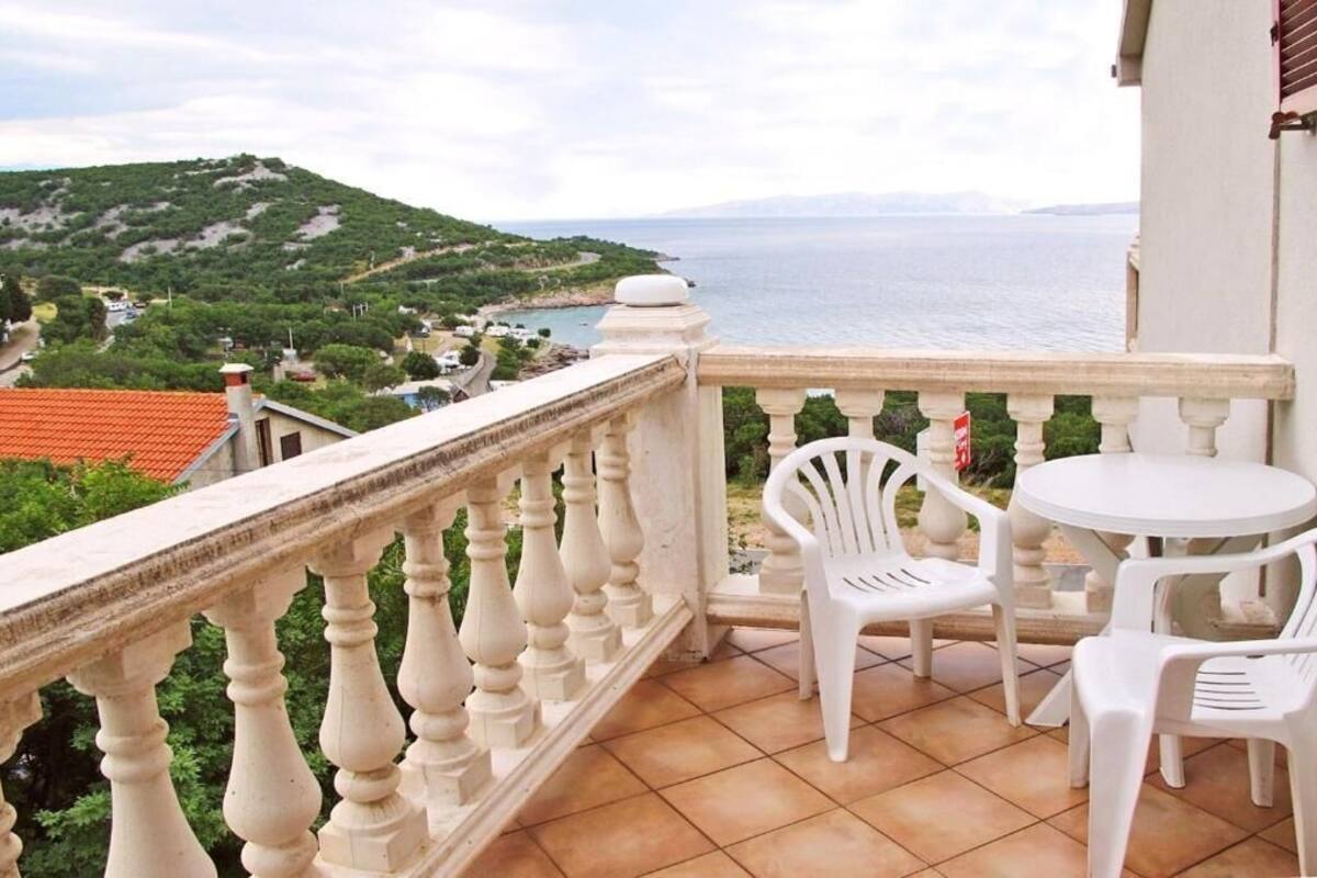 A balcony with a white balustrade, a round table with two white plastic chairs, and a view of the sea and hills in the background.