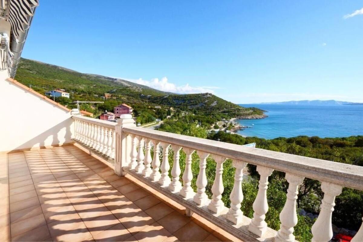A balcony with a white balustrade overlooking a coastal landscape with greenery and a clear blue sea.