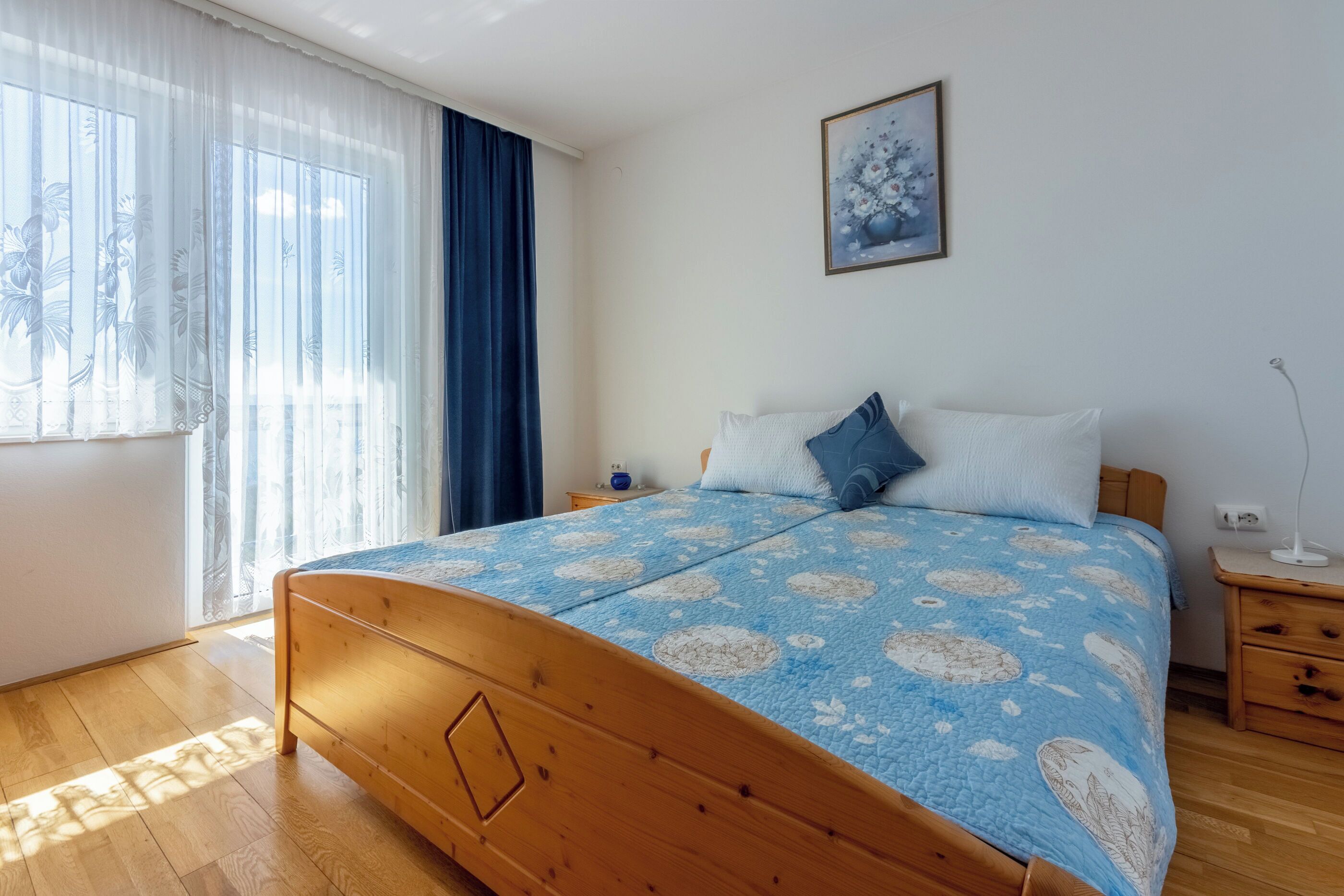Een slaapkamer met een houten bed, een blauw bloemenbedover, een nachtkastje met een lamp en een raam met dunne gordijnen.