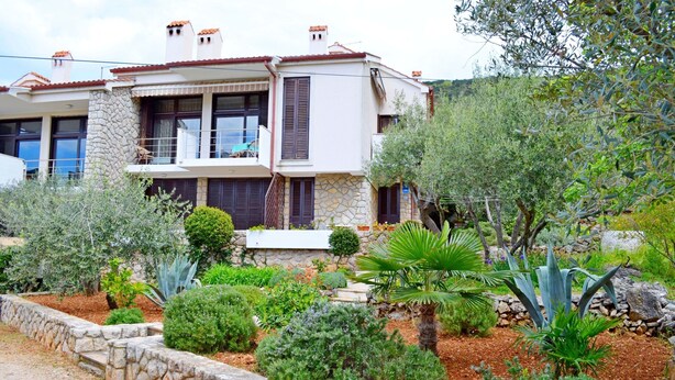 A two-story house with a stone facade, a balcony, and a well-maintained garden with various plants and a palm tree. - Apartments Marija (Cres)