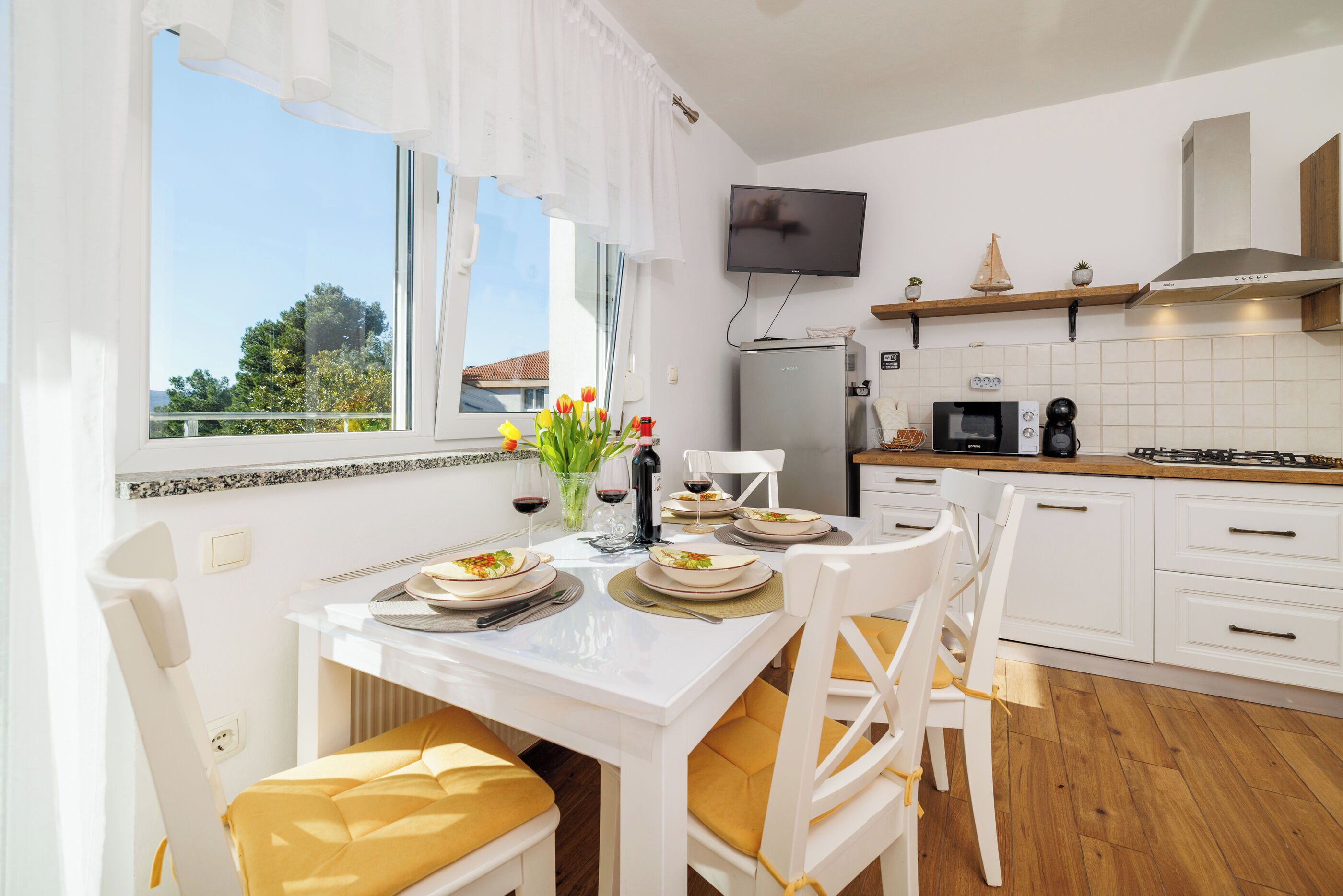 A modern kitchen with white cabinetry, a dining table set for four, and a television mounted above the counter.