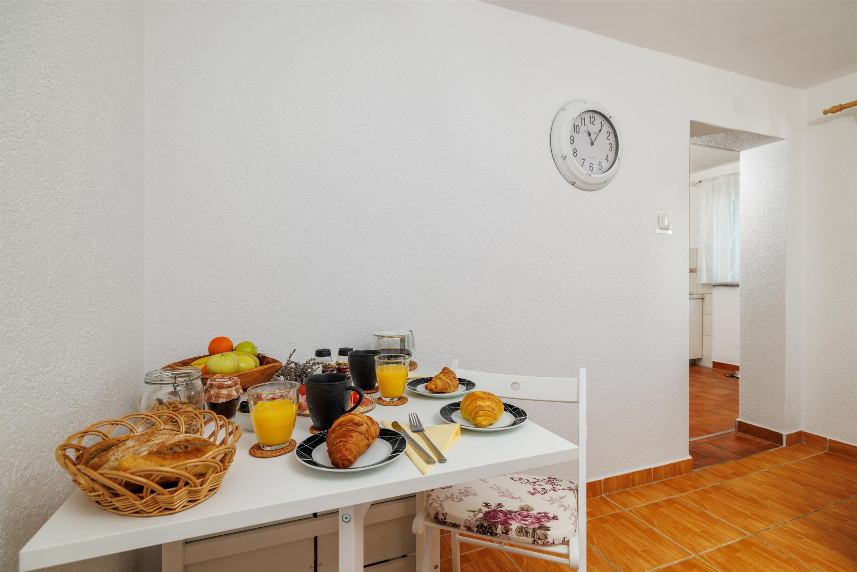 Une table de petit-déjeuner avec des croissants, des fruits et du jus, une horloge au mur et un sol carrelé.