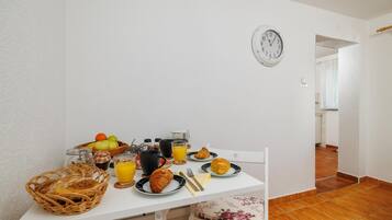 Une table de petit-déjeuner avec des croissants, des fruits et du jus, une horloge au mur et un sol carrelé.