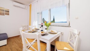 A dining area with a white table set for two, white chairs with yellow cushions, a vase with flowers, and a window with a view of trees and a building. - Apartments Iva (Crikvenica)