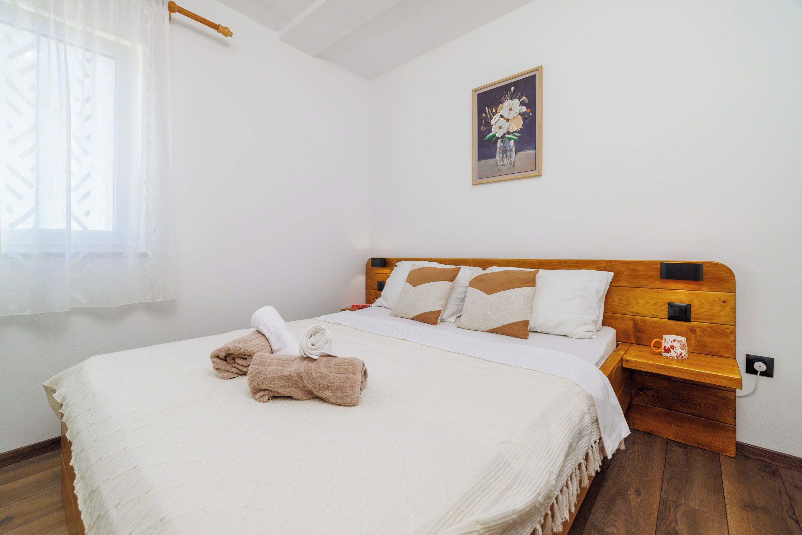 A neatly made bed with a wooden headboard, a bedside table with a mug, and a painting on the wall.