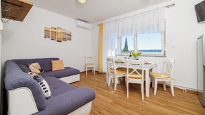 A modern living room with a grey sofa, a dining table with white chairs, and a window with curtains. - Apartments Iva (Crikvenica)
