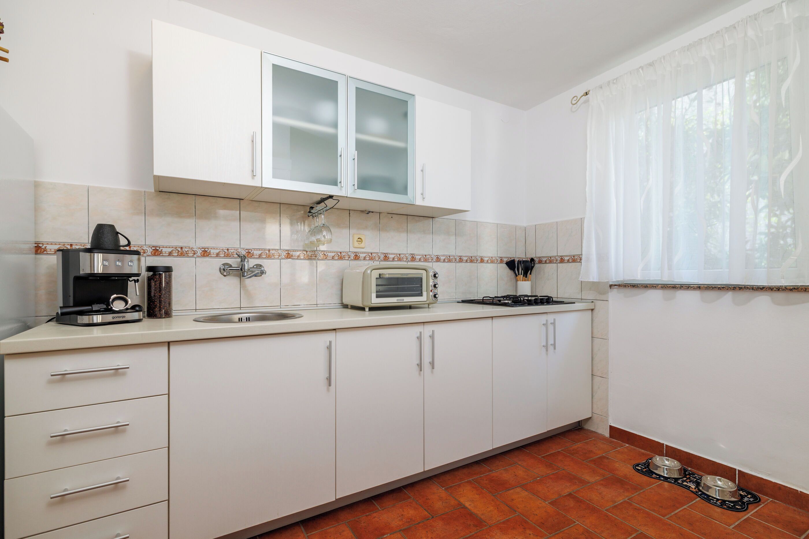 A kitchen with white cabinets, a toaster, a coffee maker, and a window with curtains.