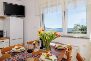 A dining area with a table set for two, a television above, and a window with a view of the sea.