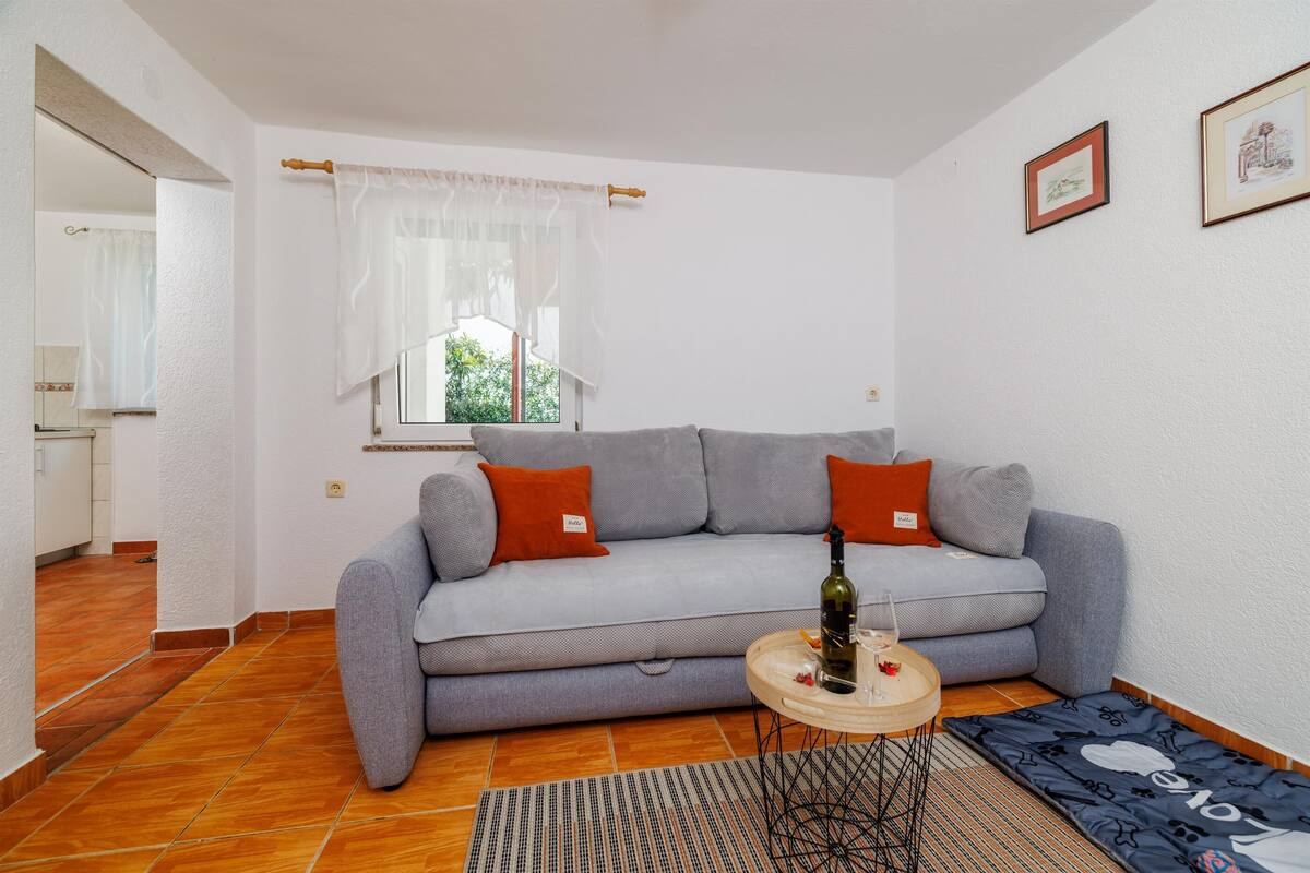 A living room with a grey sofa, two orange pillows, a small round table with a bottle and glass, and a patterned rug.