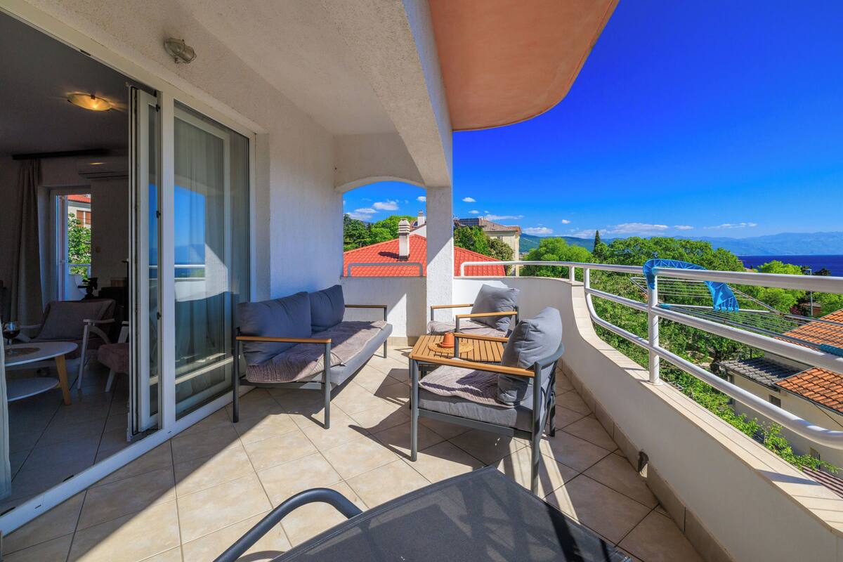 A balcony with a table and chairs, a sliding glass door, and a view of a red-roofed building and greenery.