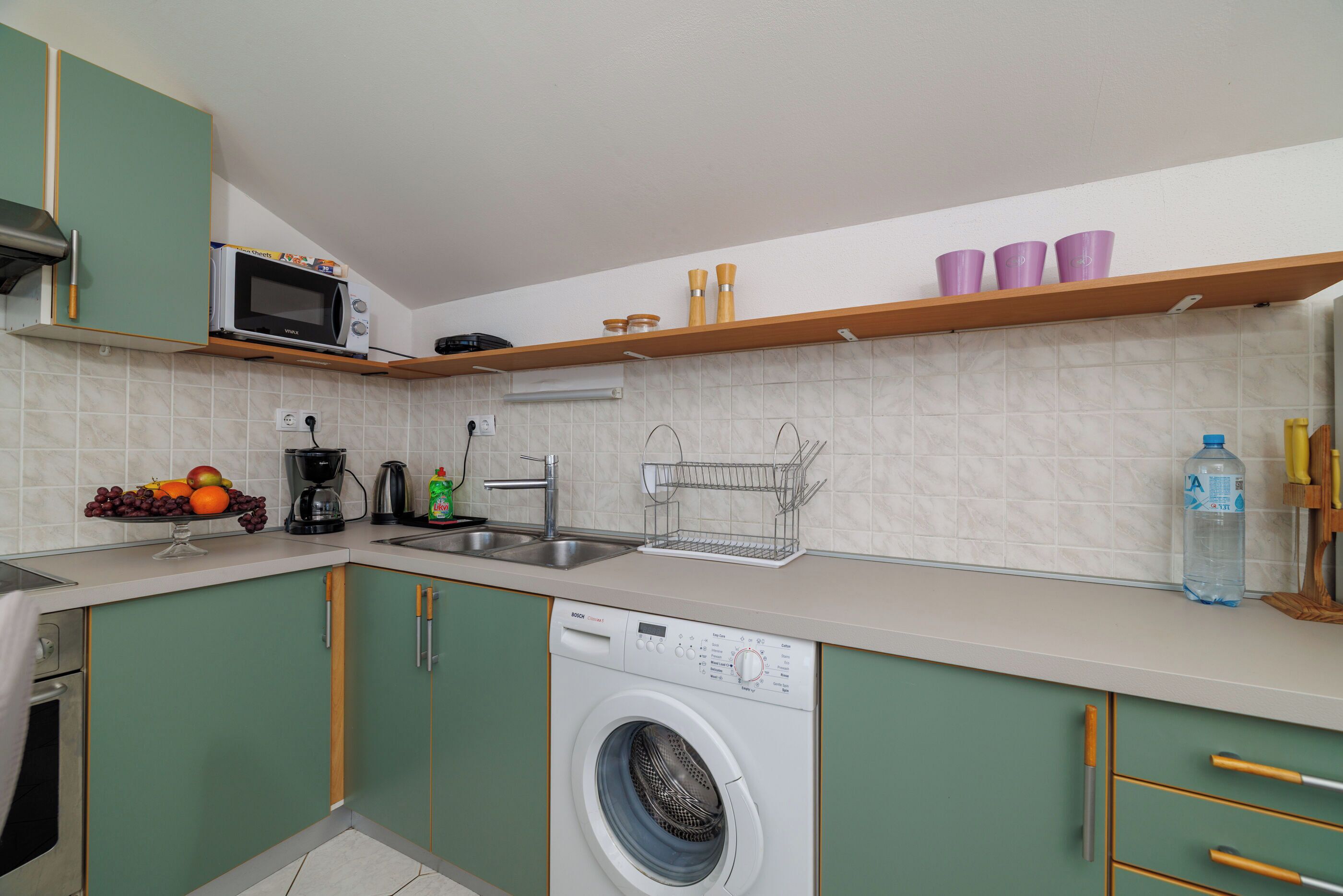 A compact kitchen with a washing machine, a microwave, and a fruit bowl on the counter.