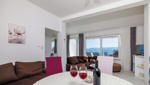 A modern living room with a brown sofa, a white round table with two wine glasses and a bottle, and a view of the sea through large windows.
