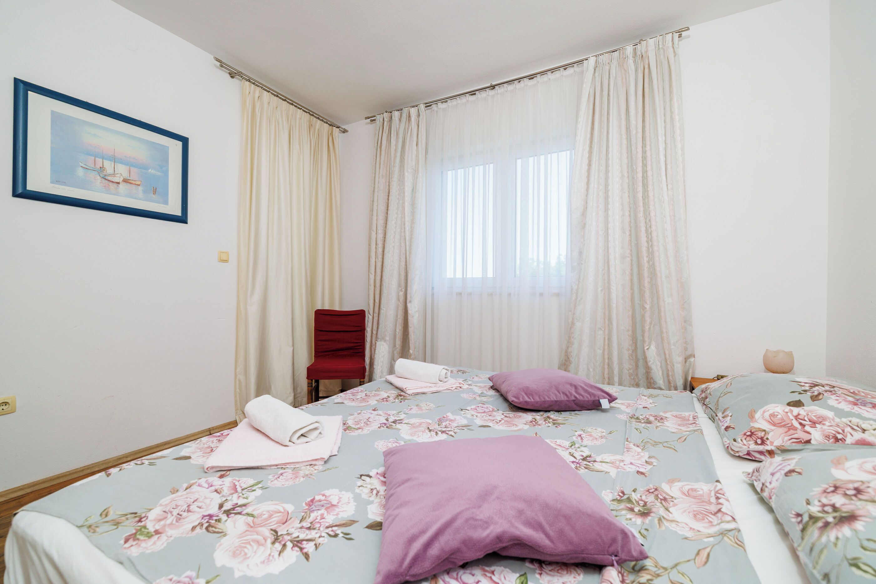A bedroom with two beds, floral bedding, a red chair, and a painting on the wall.