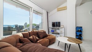 A modern living room with a large brown sofa, a white coffee table, a flat-screen TV, and a view of the sea through a sliding glass door.
