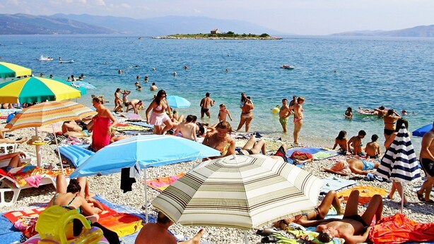 A crowded beach with people sunbathing and swimming, surrounded by umbrellas and beach chairs.