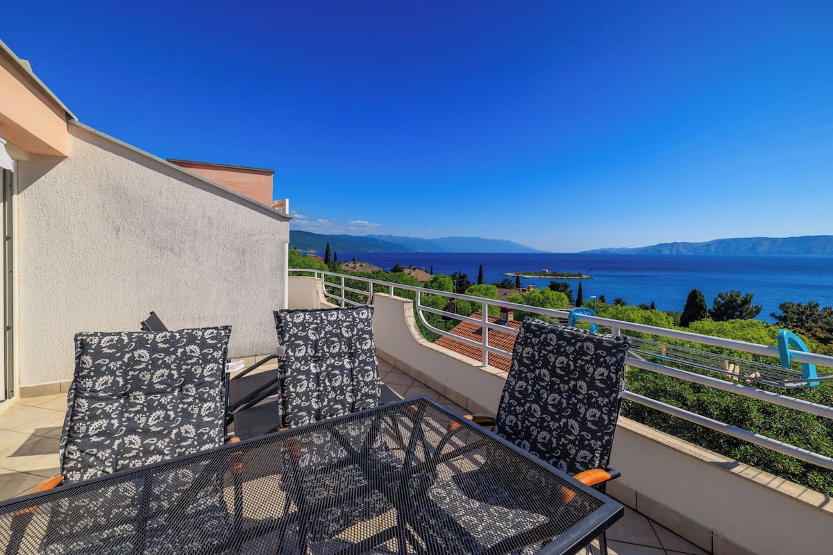A balcony with outdoor furniture, a clear view of the sea, and a mountainous landscape in the distance.