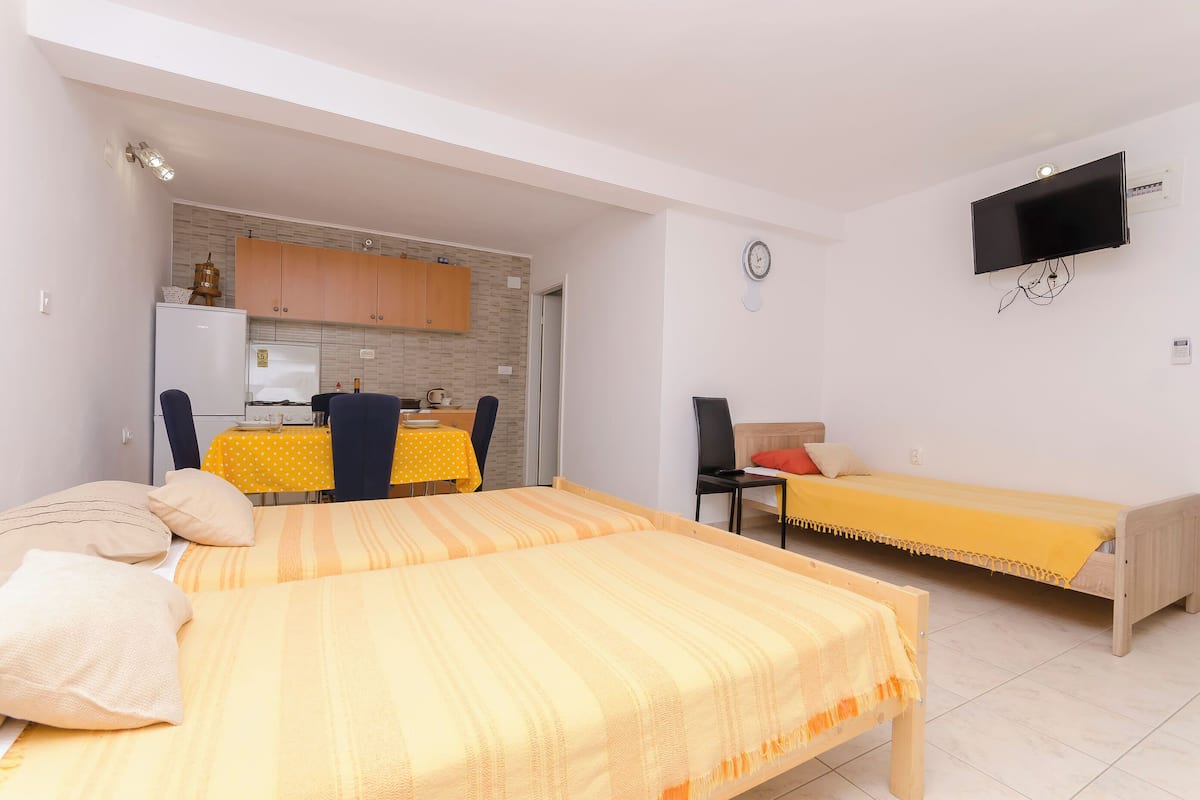 A compact room with two beds, a TV, a clock, and a kitchenette in the background.