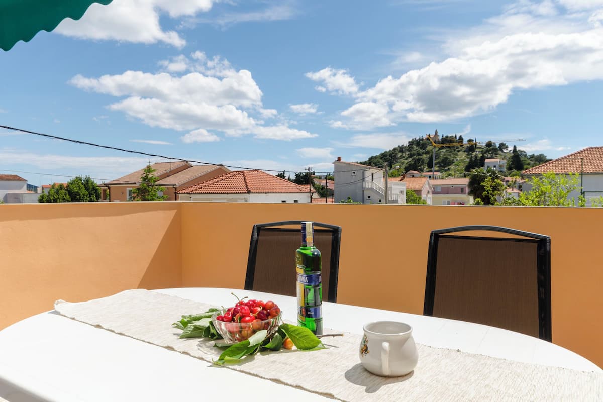 A table with a bowl of cherries, a bottle, and a cup on a balcony overlooking a residential area with houses and a hill in the background.