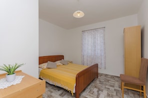 A bedroom with a wooden bed, a wooden wardrobe, a wooden chair, a small table with a plant, and a window with floral curtains. - Apartments Mila II (Tribunj)