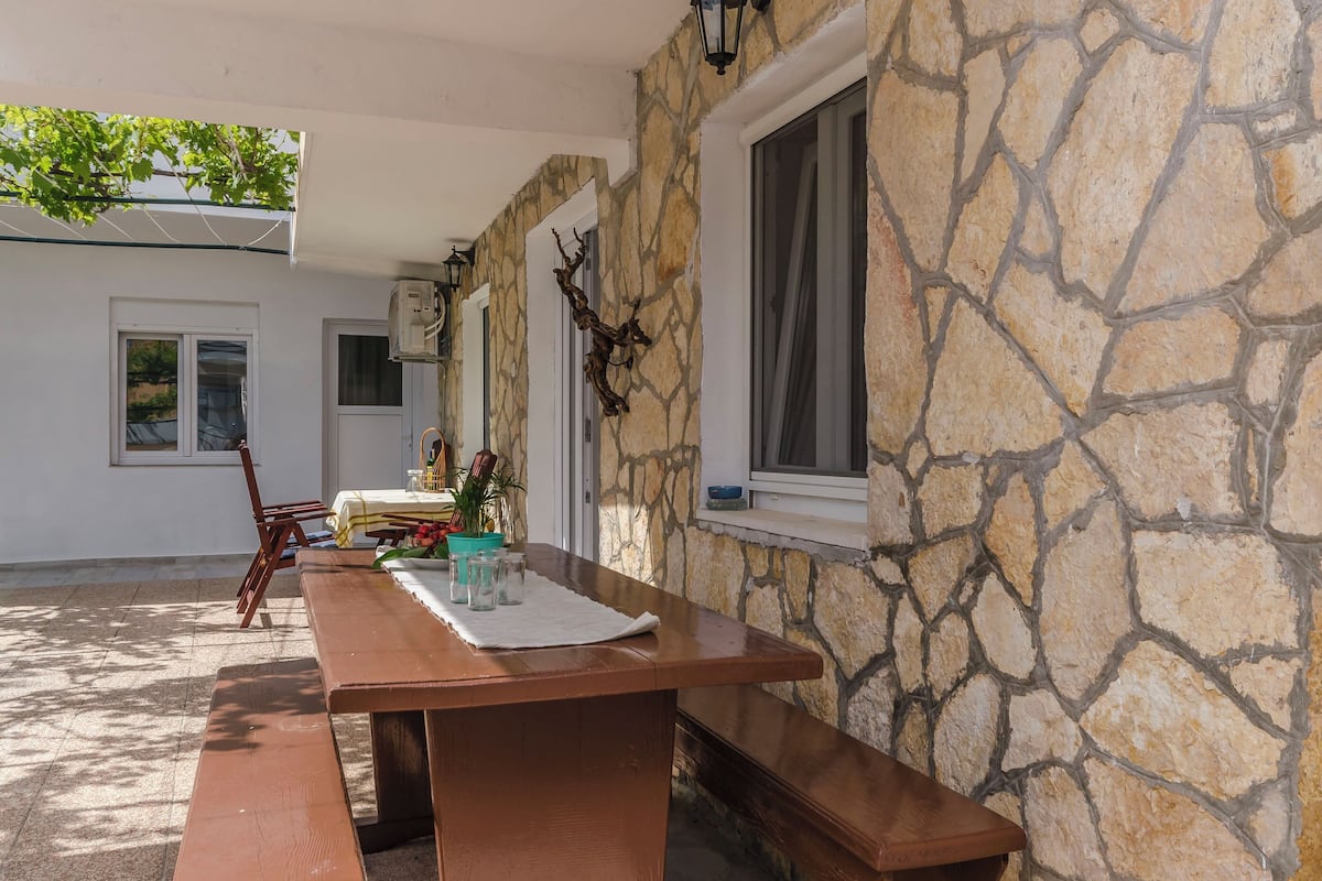 An outdoor patio with a wooden table and benches, stone walls, and a window with curtains.