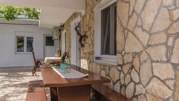 Une terrasse extérieure avec une table et des bancs en bois, des murs en pierre et une fenêtre avec des rideaux.