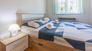 A neatly made bed with a blue and white quilt, a bedside lamp, and a window with sheer curtains. - Apartments Ivana (Crikvenica)