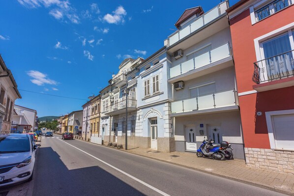 A street with parked cars and a scooter, buildings with balconies, and a clear blue sky.