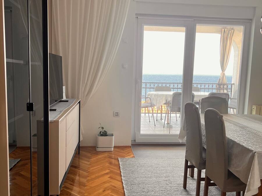 A modern living room with a wooden floor, a glass door leading to a balcony with a view of the sea, a dining area with a table and chairs, and a TV on a stand.