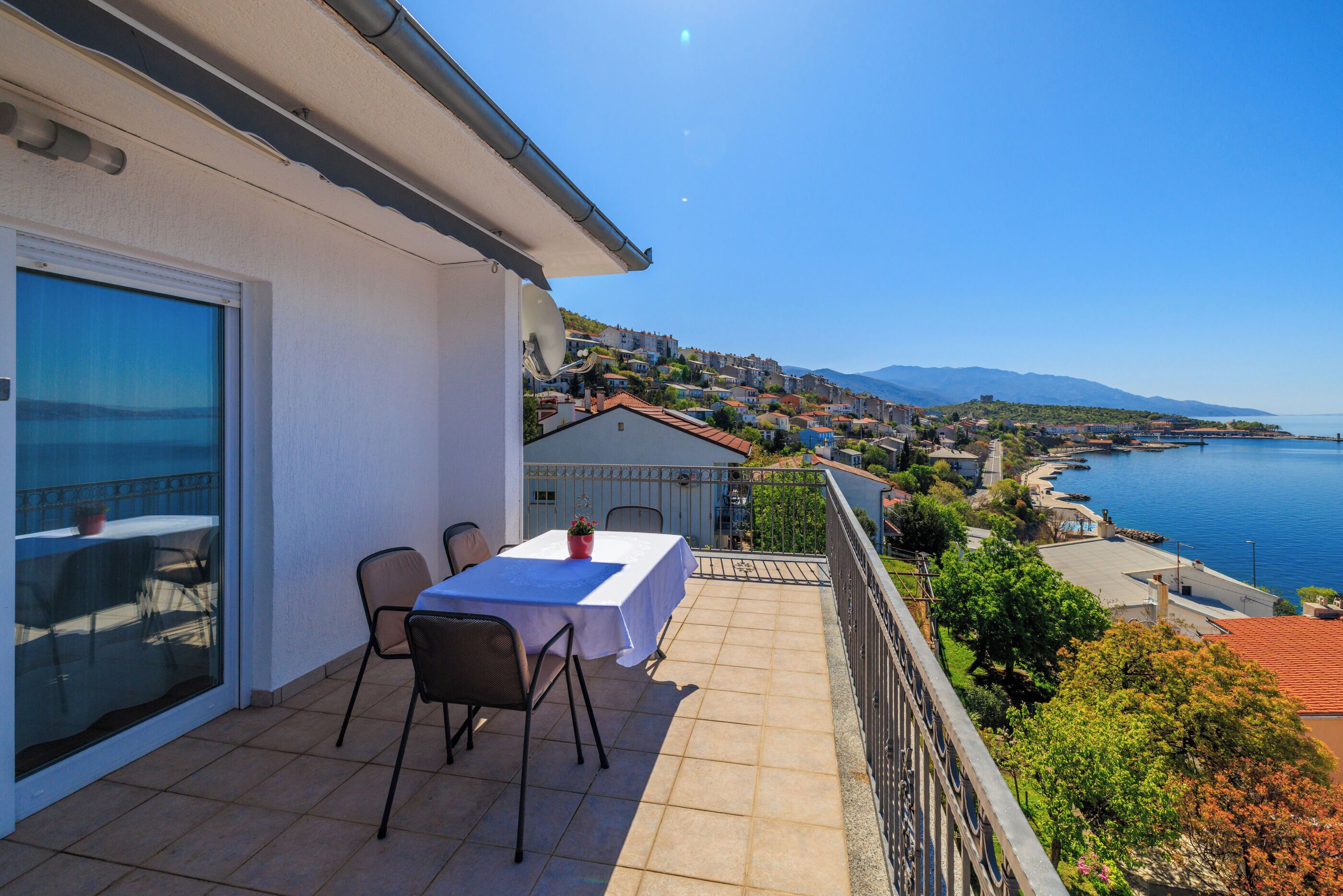 A balcony with a table and chairs, overlooking a coastal town and the sea.