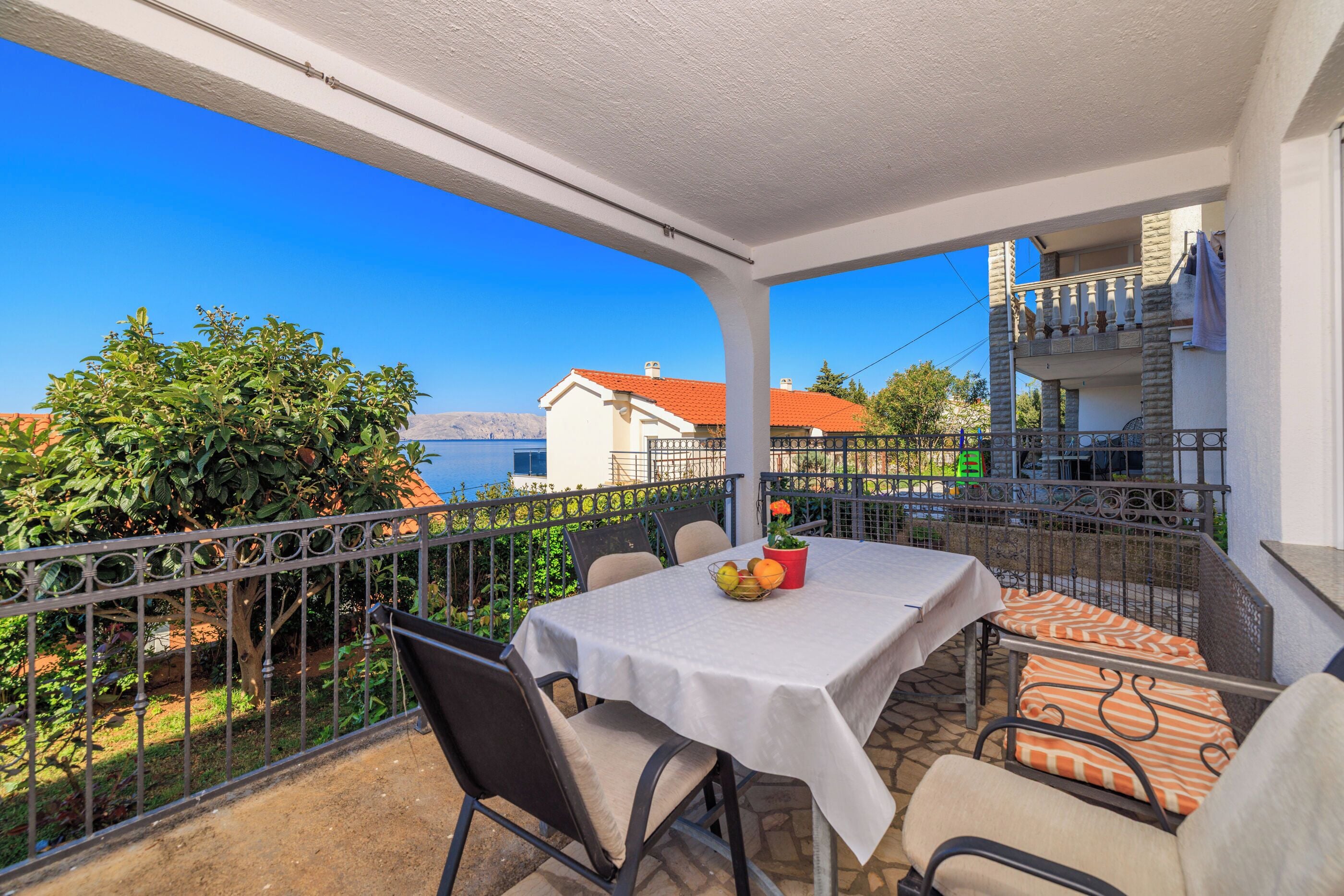A balcony with a table set for two, outdoor seating, and a view of the sea and buildings.