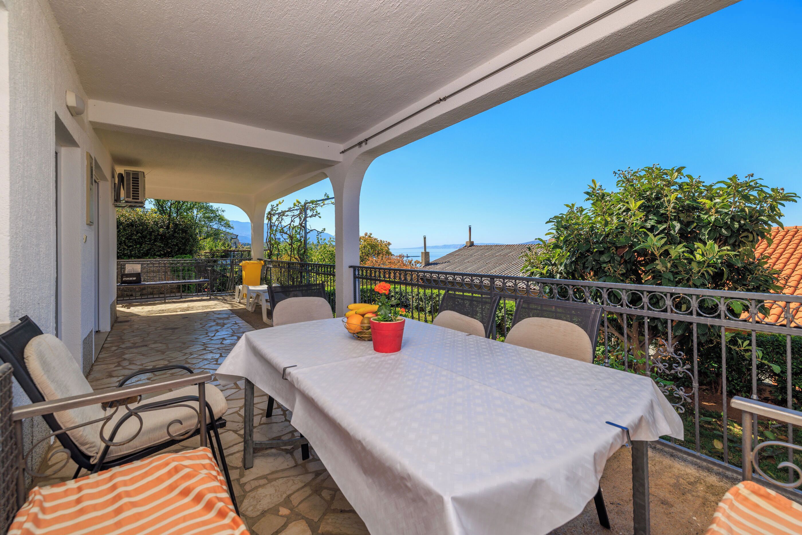 Une terrasse couverte avec une table et des chaises, un pot de fleurs rouge, et une vue sur la mer et les collines.