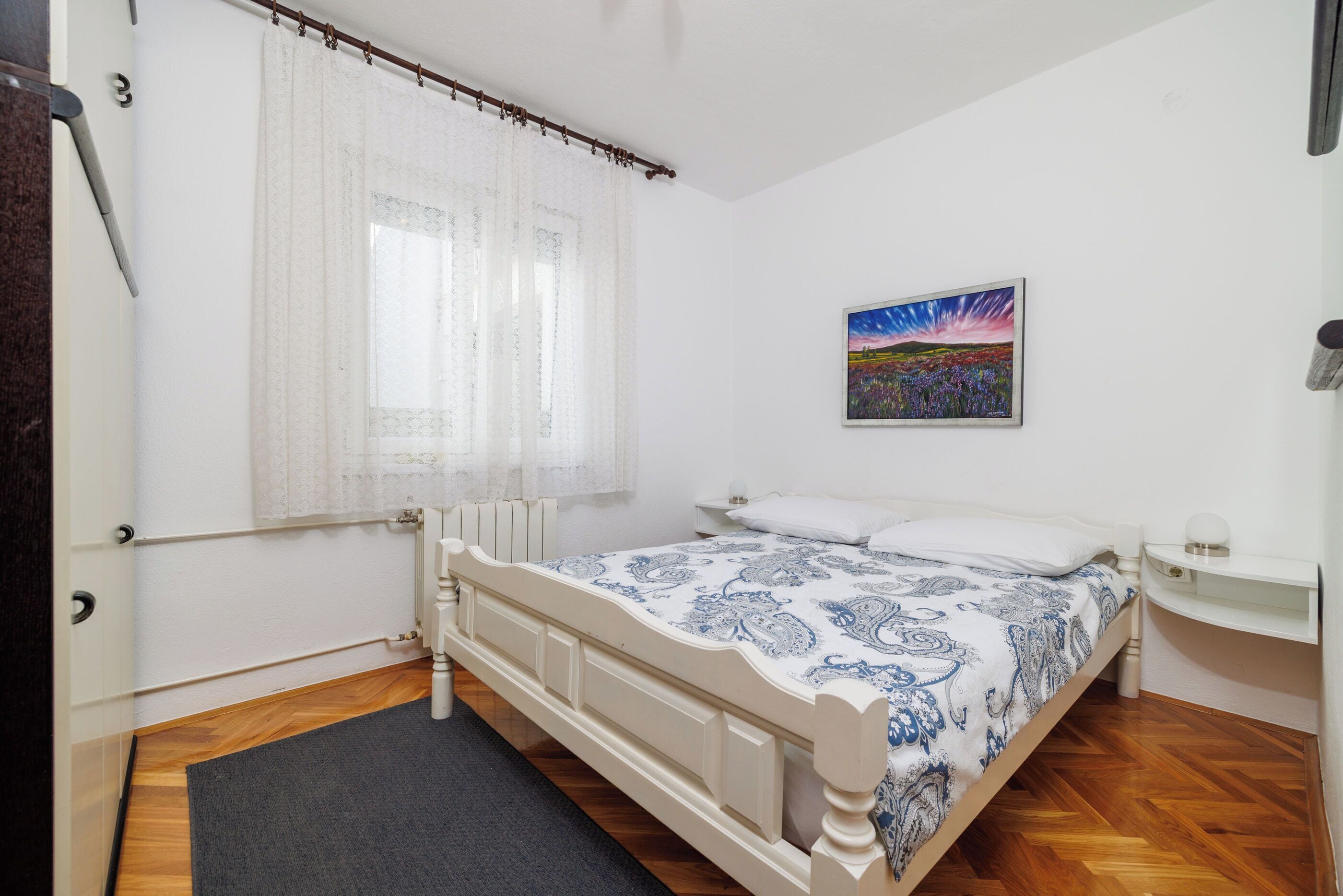 Un dormitorio con cama, mesita de noche, un cuadro y ventana con cortinas.