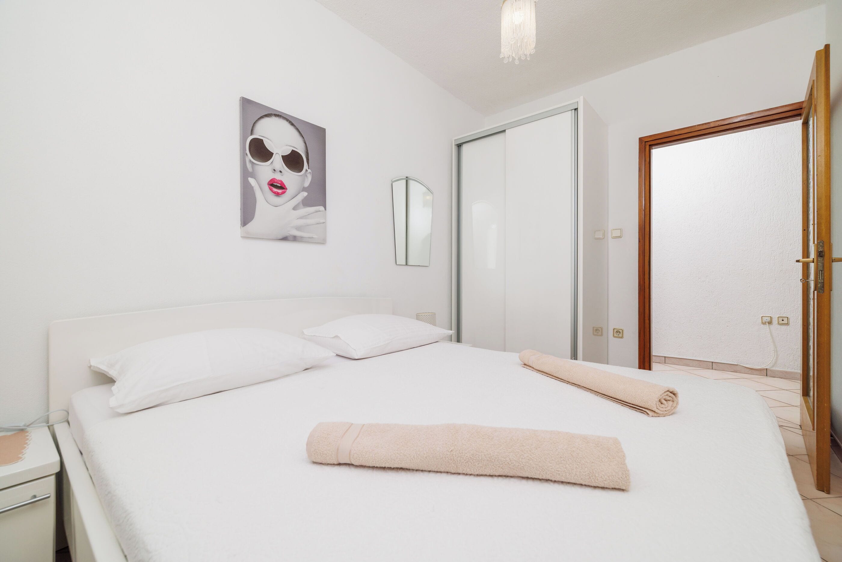 A neatly made bed with white linens and a rolled towel, a white nightstand, a mirror, and a door to the right.
