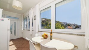 A kitchen with a dining table, chairs, a bowl of fruit, a wine bottle, and a view of buildings through a window.