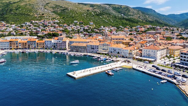A coastal town with a marina, buildings with red-tiled roofs, and a hilly backdrop.