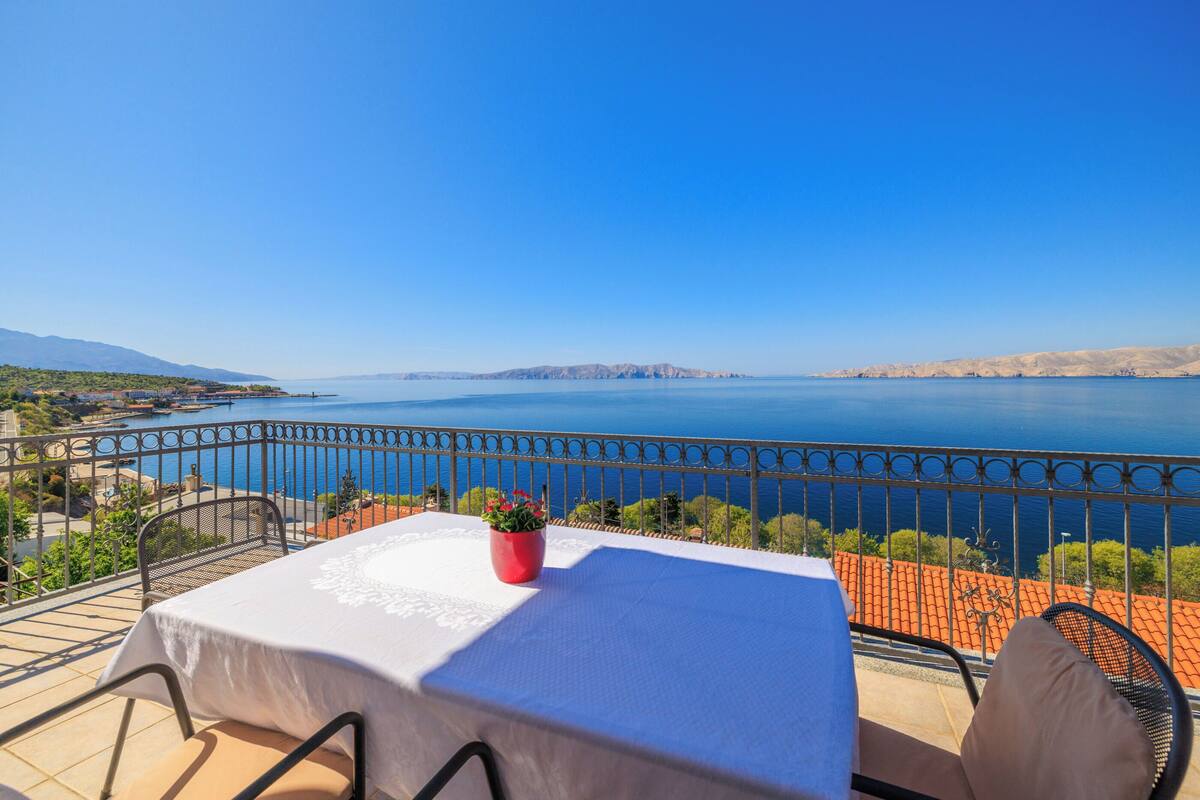 Una terraza con una mesa y sillas, con vista a una gran masa de agua y montañas distantes.