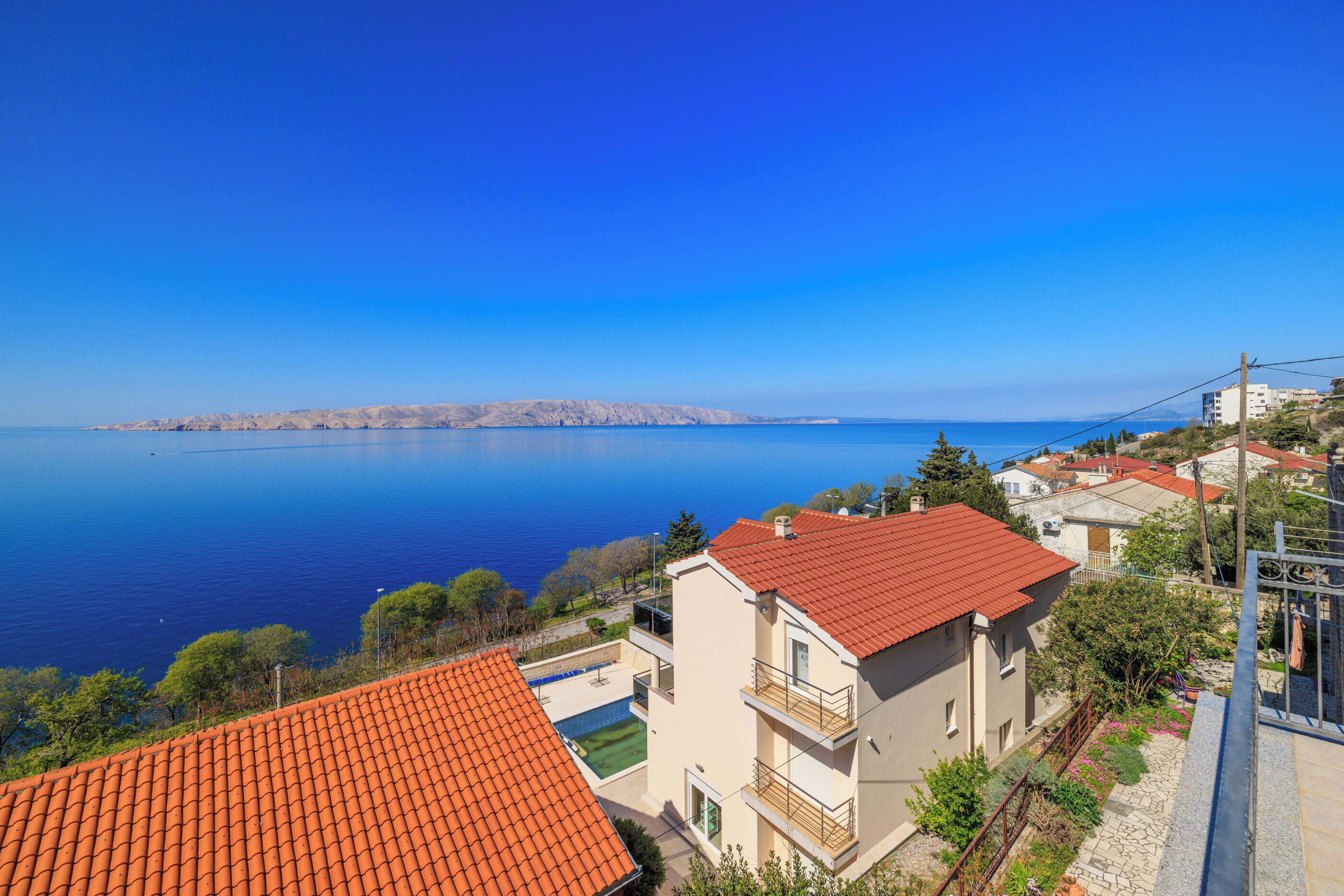 A coastal residential area with a clear view of the sea, a building with a red-tiled roof, and a balcony.