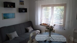 Een gezellige woonkamer met een grijze bank, een kleine tafel met een kanttafelkleed en een raam met witte gordijnen.
