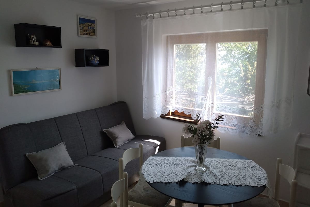 A cozy living room with a grey sofa, a small table with a lace tablecloth, and a window with white curtains.