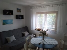 Una sala de estar acogedora con un sofá gris, una mesita con mantel de encaje y una ventana con cortinas blancas.
