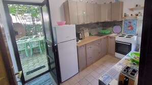 Een kleine keuken met een koelkast, fornuis en wastafel. Kastjes en een klein aanrecht met diverse spullen.
