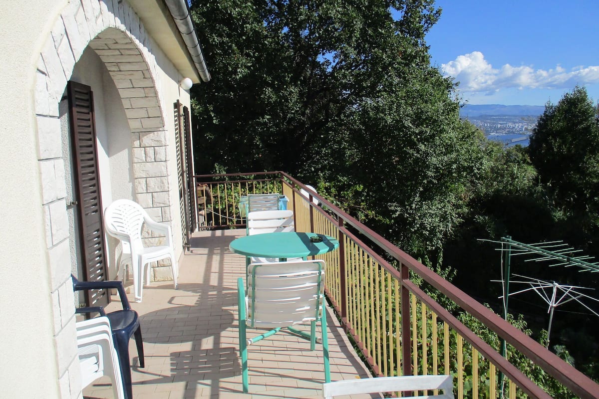 A balcony with outdoor furniture, a view of trees and a distant cityscape.