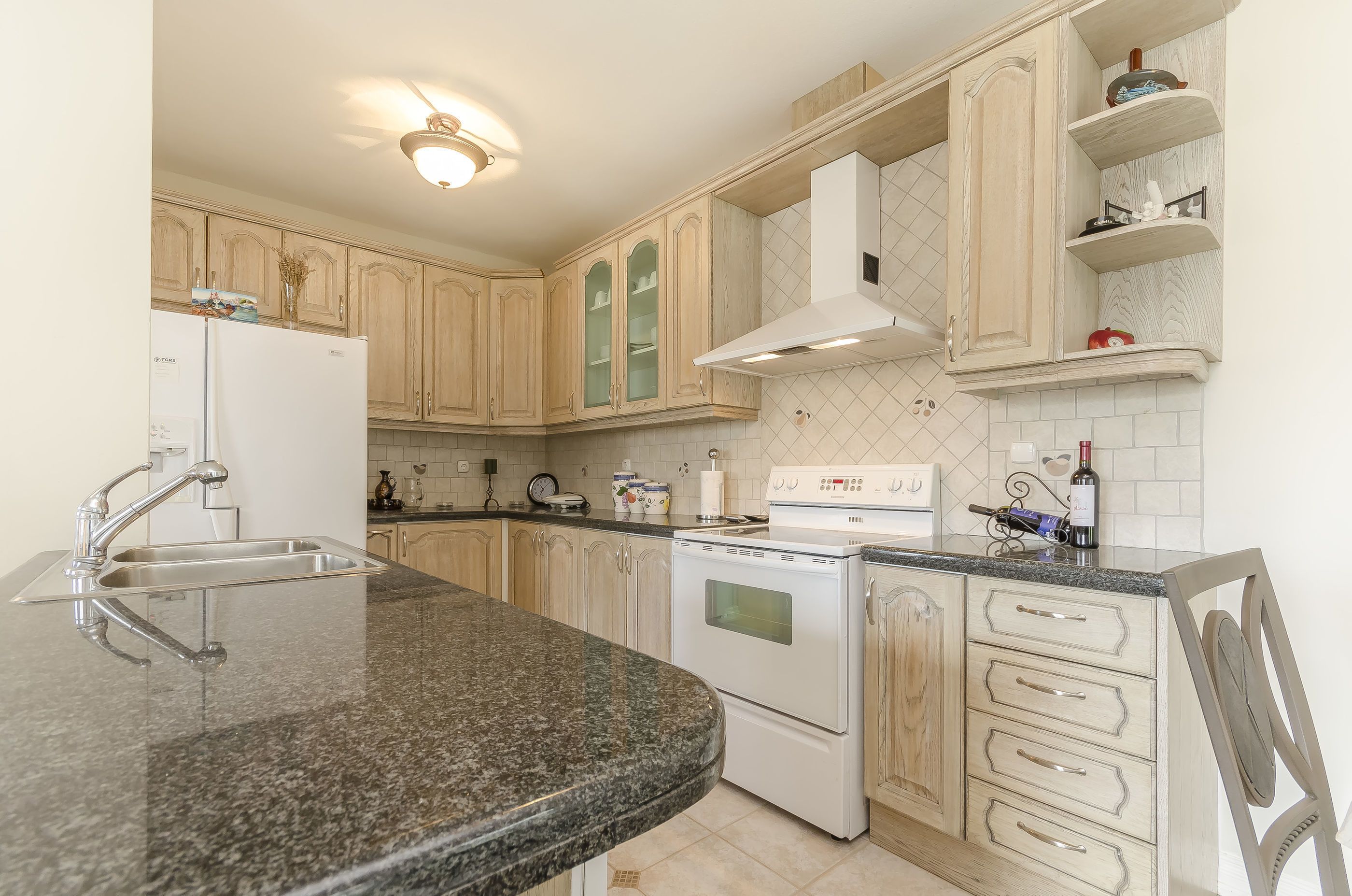 A kitchen with a granite countertop, stainless steel sink, white appliances, and wooden cabinets.