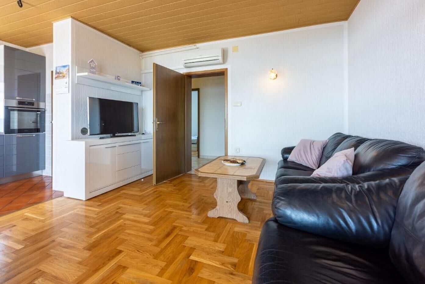 Un saló modern amb un sofà de pell negra, una taula de centre de fusta i una zona de cuina amb televisió integrada i forn.