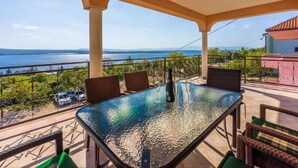Une table à plateau en verre, quatre chaises, une bouteille et des verres sont disposés sur un balcon donnant sur un vignoble et la mer.
