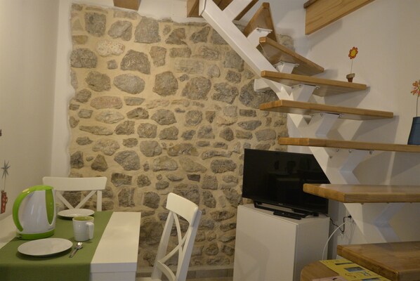 A room with a stone wall, a wooden staircase, a television, a table with chairs, and a green tablecloth. - Apartment Oliva (Cres)
