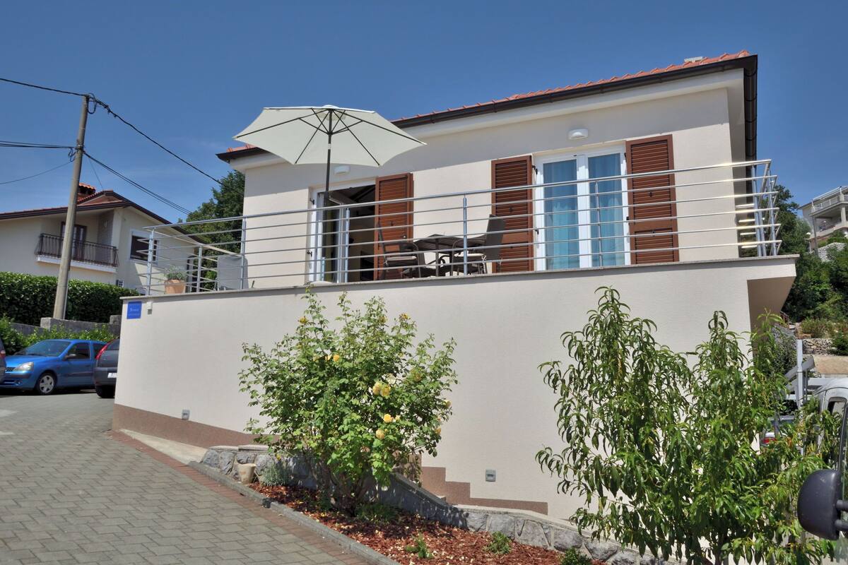 Una moderna casa a due piani con un balcone, un patio con mobili da esterno e un giardino con piante.