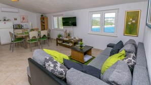 Ein modernes Wohnzimmer mit grauem Sofa, grünem und gemusterten Kissen, einem Holztisch und einem Flachbildfernseher. Es gibt einen Essbereich mit Tisch und Stühlen sowie eine Küchenzeile mit Kühlschrank, Mikrowelle und Spüle.