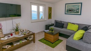 Ein Wohnzimmer mit grauem Sofa, einem Holztisch, einem an der Wand montierten Fernseher, einem Fenster mit Meerblick und einem Bild an der Wand.