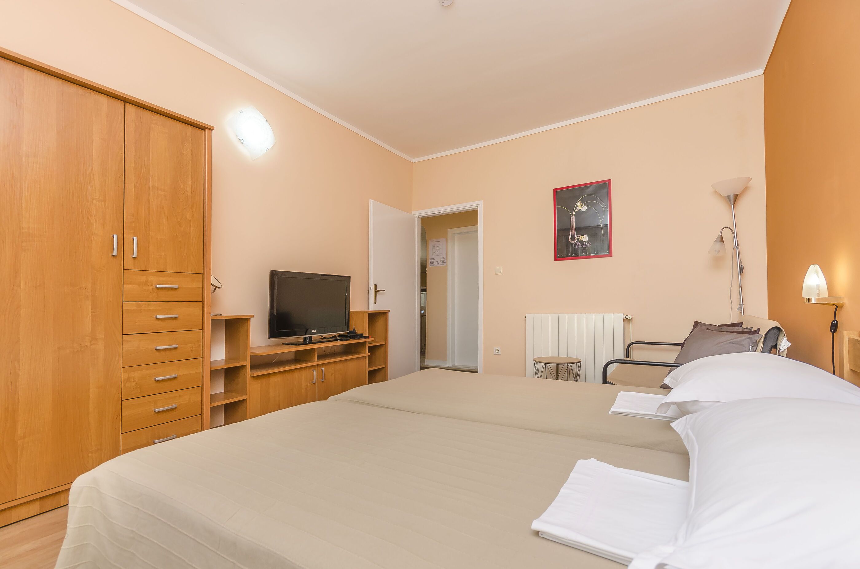 A bedroom with a bed, wooden wardrobe, TV, bedside table, lamp, and a door leading to another room.